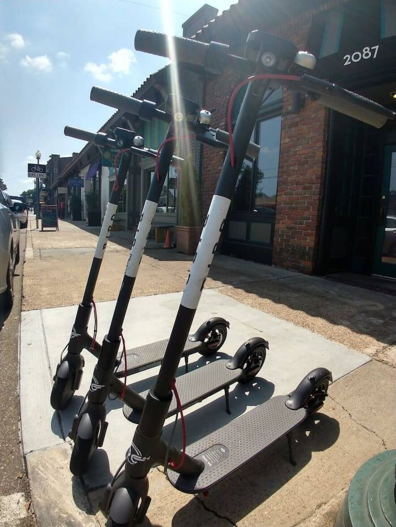A row of electric scooters parked on a sidewalk in front of a building with the number 2087.