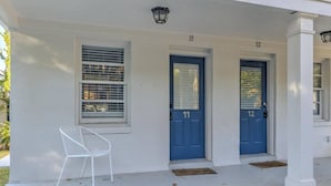 Un edificio blanco con dos puertas azules, identificadas con los números 11 y 12, una silla blanca y una pequeña zona de porche.