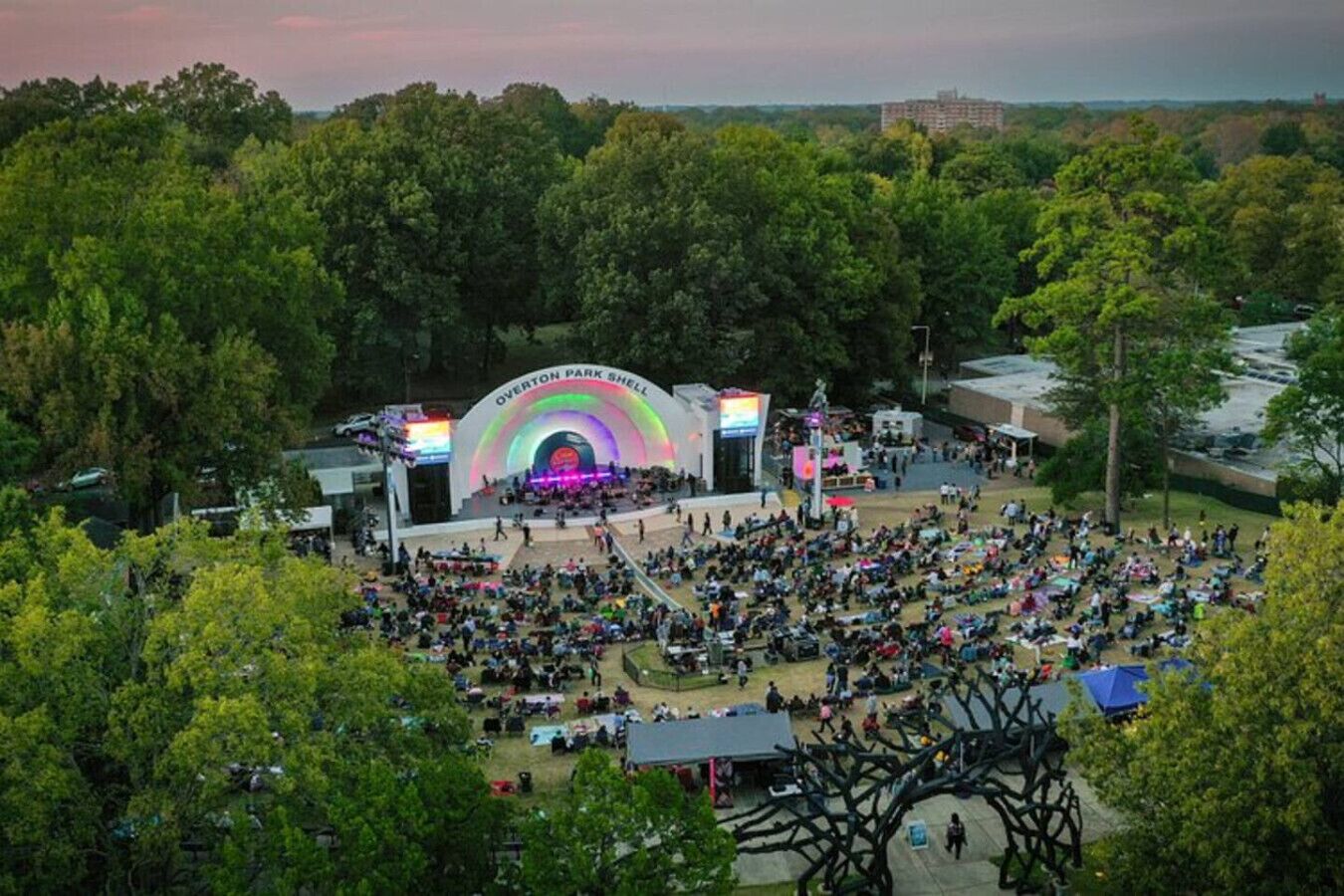 A large outdoor amphitheater with a rainbow arch and a stage, surrounded by trees and a crowd of people.