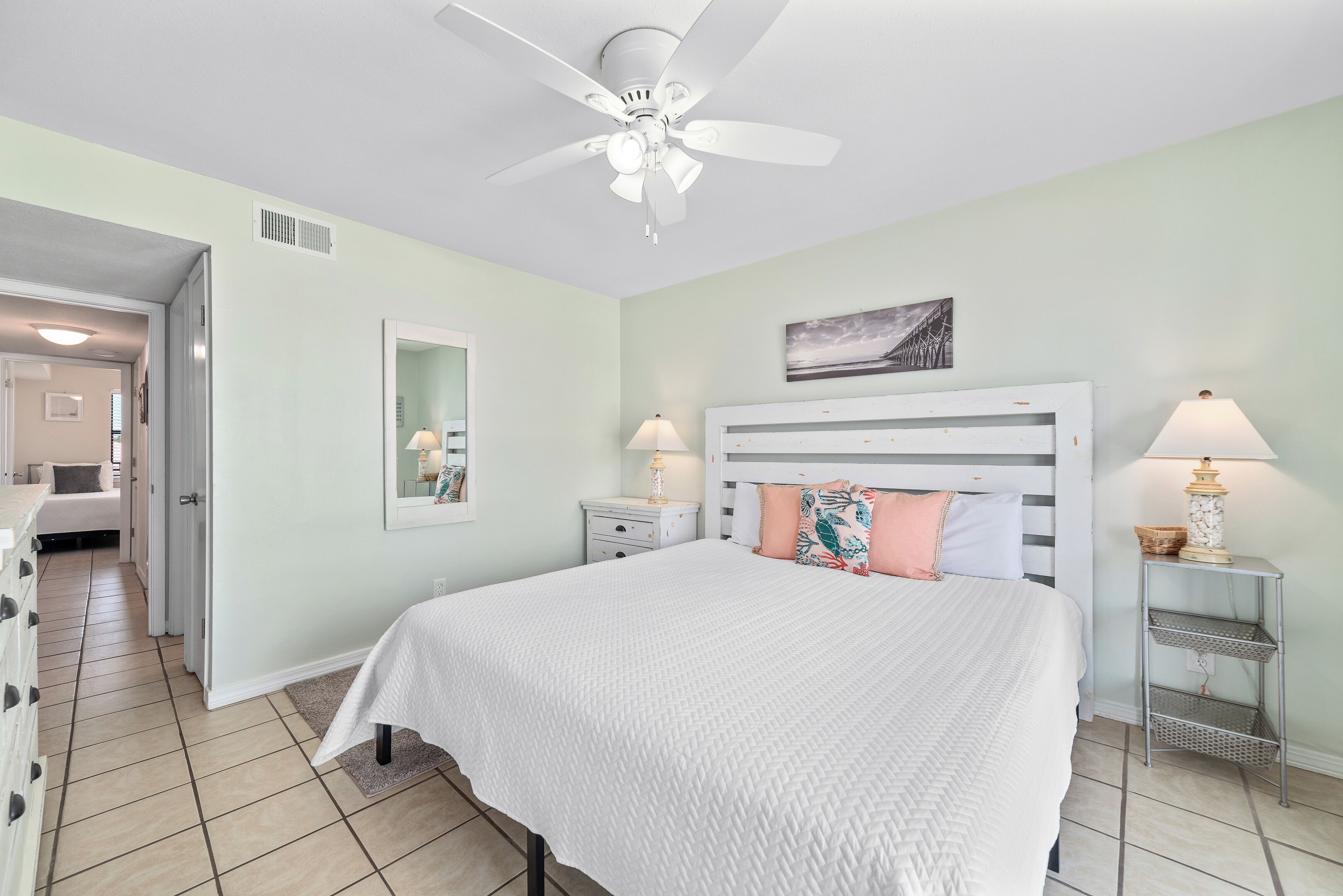 A bedroom with a bed, a ceiling fan, a nightstand with a lamp, a bedside table with a lamp, and a wall-mounted shelf.