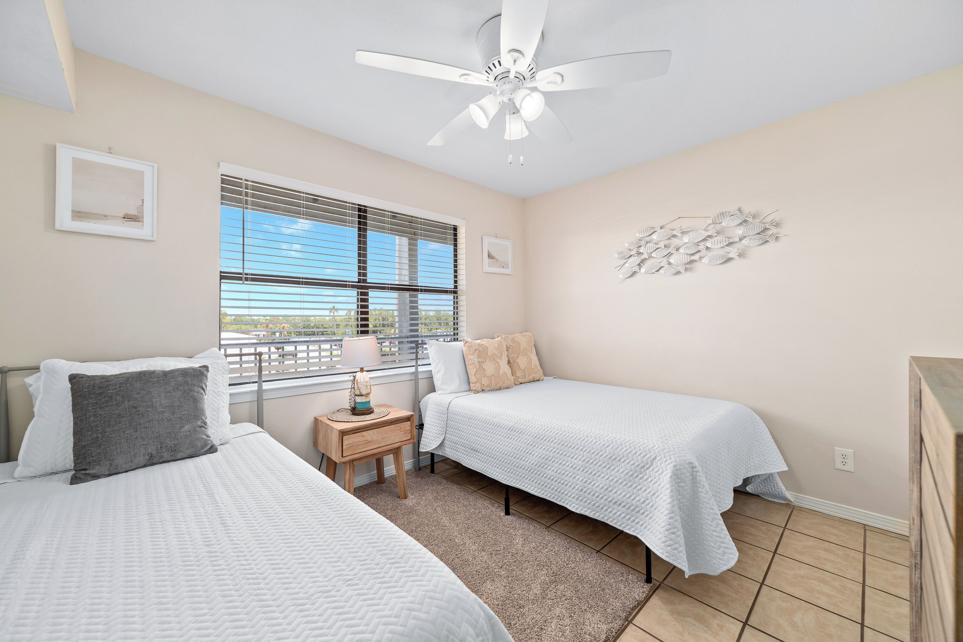 A bedroom with two beds, a ceiling fan, a window with blinds, and a small table with a lamp.
