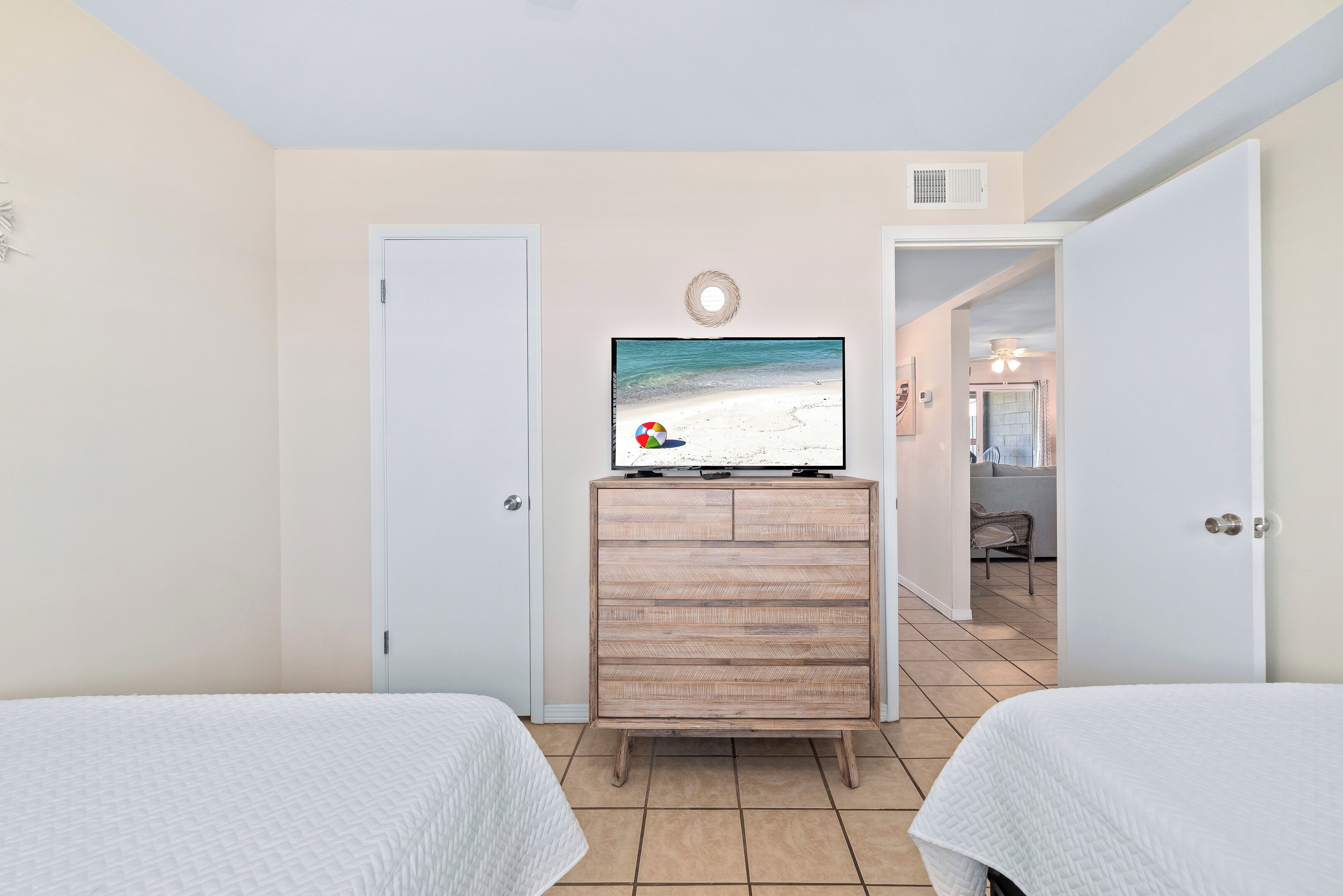 Ein Hotelzimmer mit zwei Betten, einem hölzernen Schminktisch, einem Fernseher, der eine Strandansicht zeigt, und einer Tür zu einem anderen Raum.