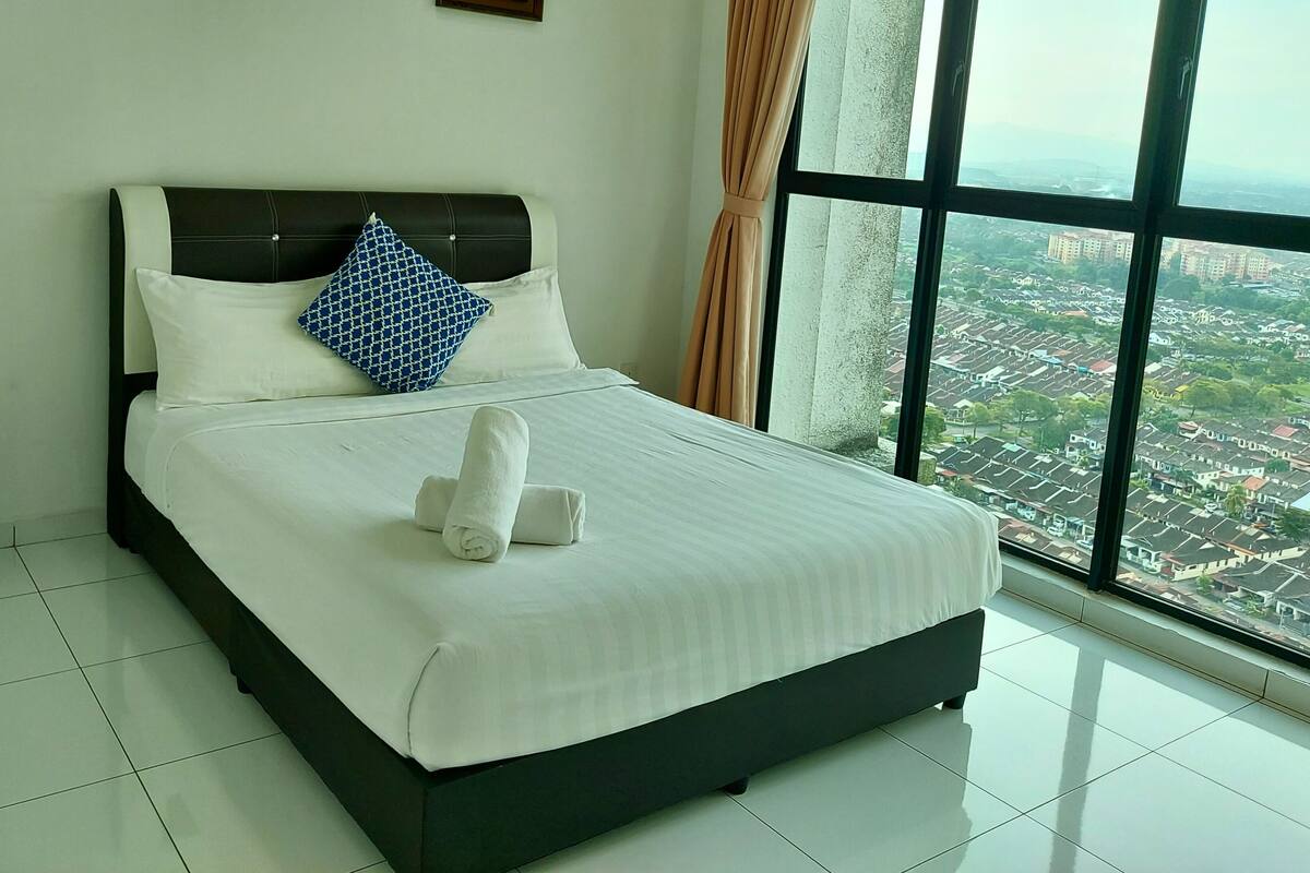 A hotel room with a large bed, a nightstand, and a view of a cityscape through a large window.