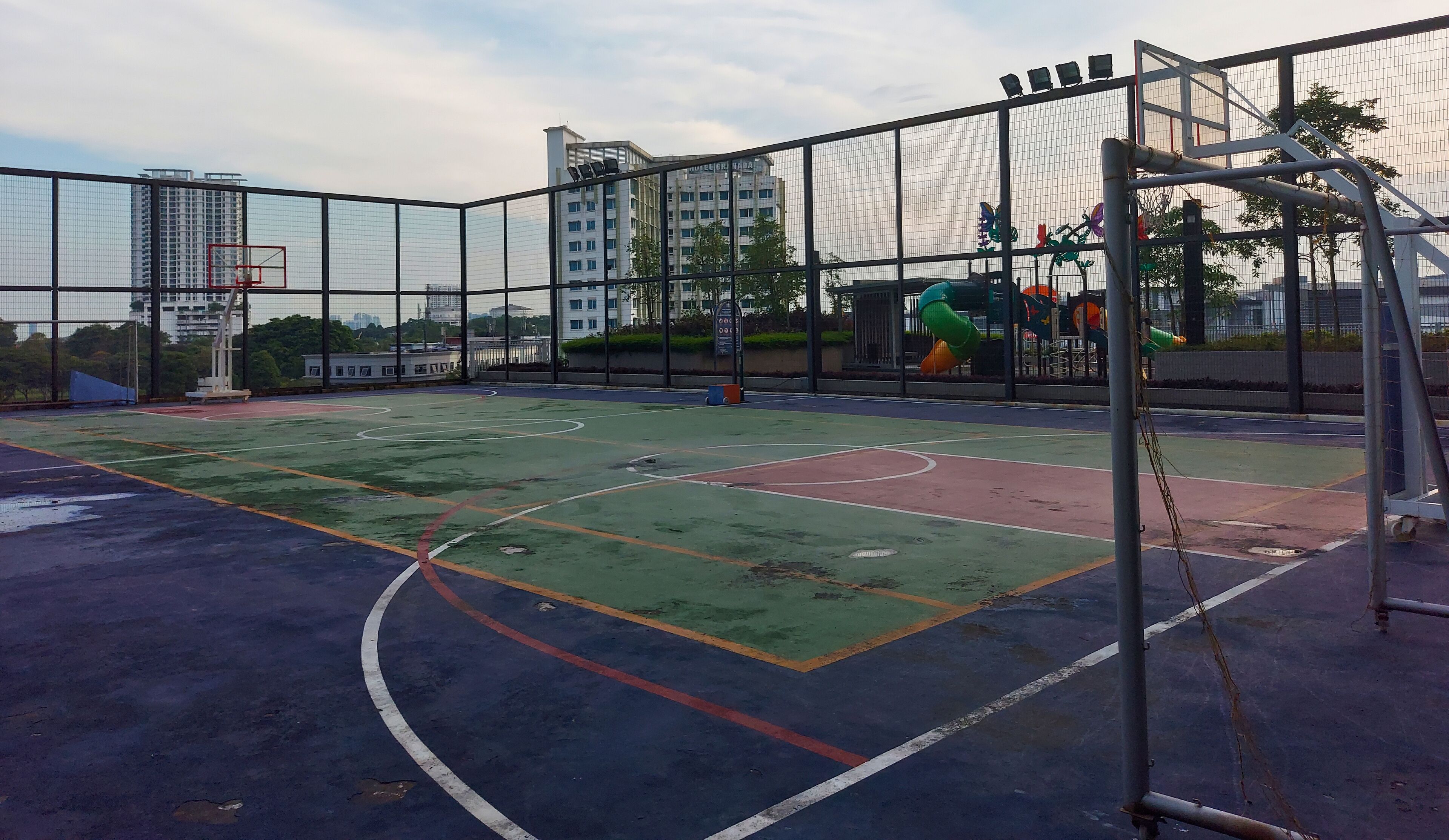 An outdoor basketball court with a netted fence, surrounded by buildings and trees.