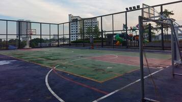 An outdoor basketball court with a netted fence, surrounded by buildings and trees.