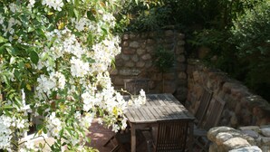 An outdoor patio with a stone wall, wooden table, and chairs, surrounded by lush greenery and white flowers.