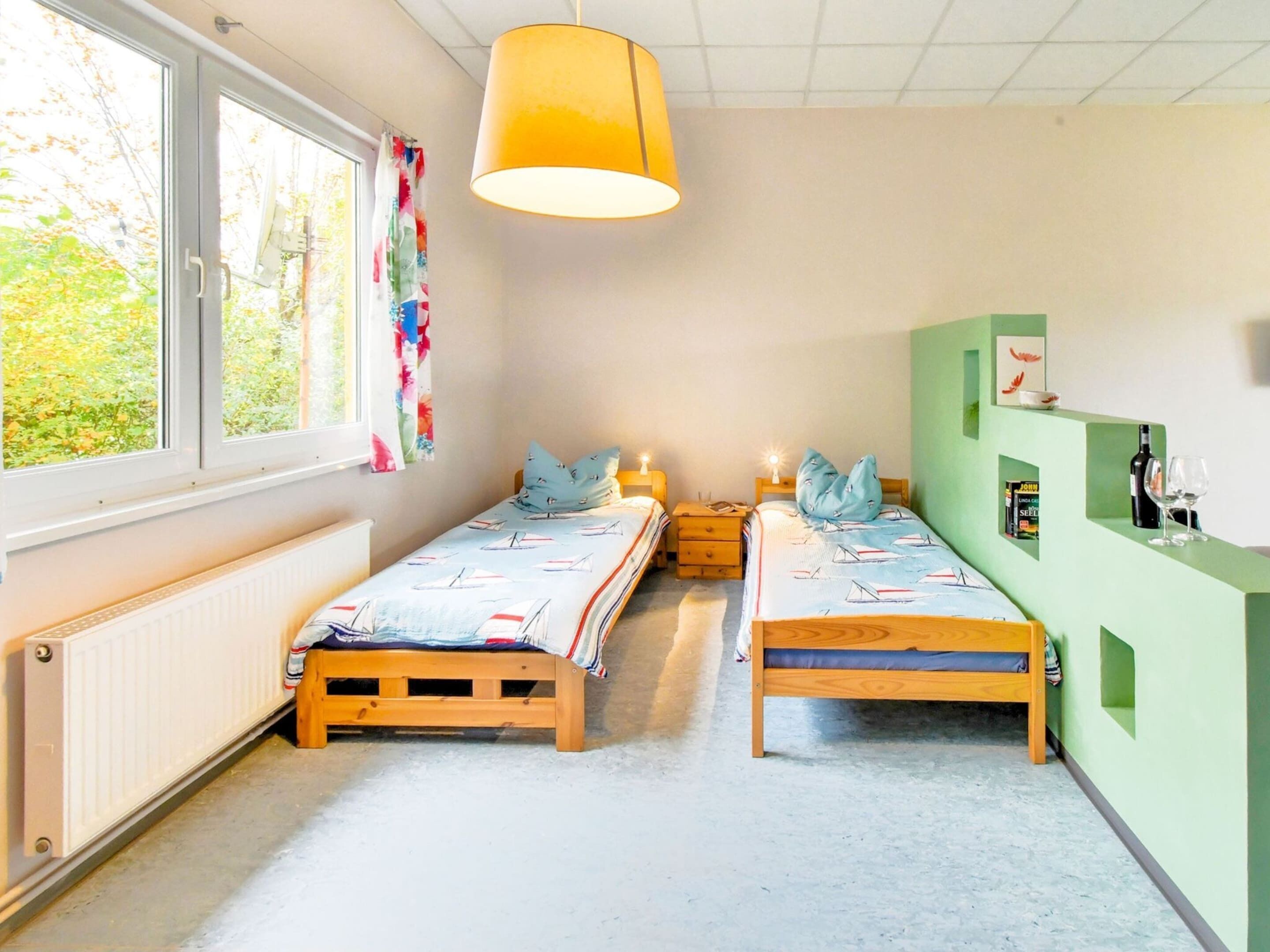 A room with two wooden beds, a large window with curtains, a hanging lamp, and a green wall with a shelf.