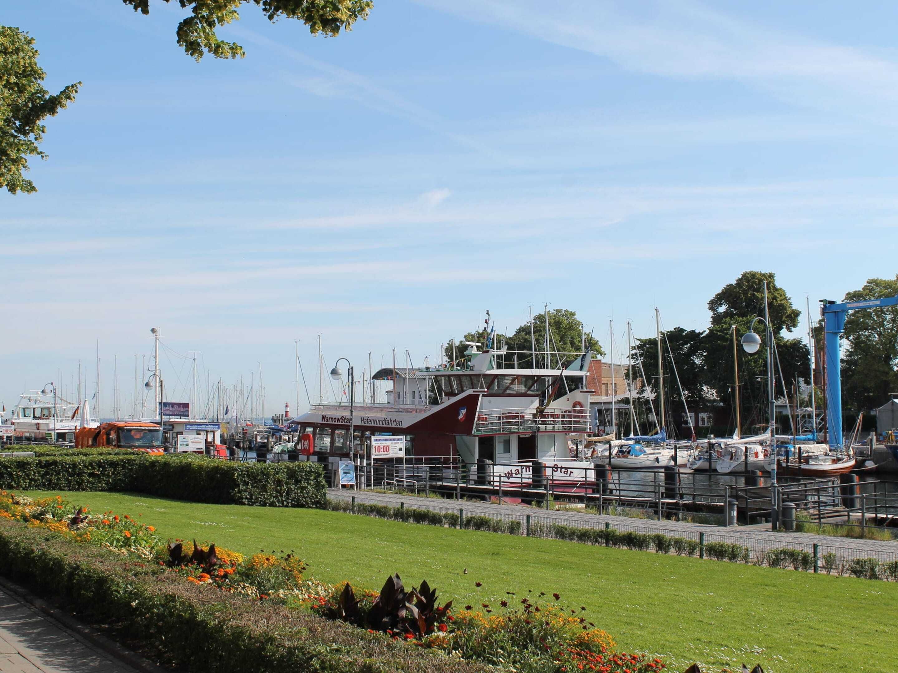 Un puerto deportivo con barcos amarrados, un edificio con el rótulo 'WarnowStar' y una zona ajardinada bien cuidada con flores.