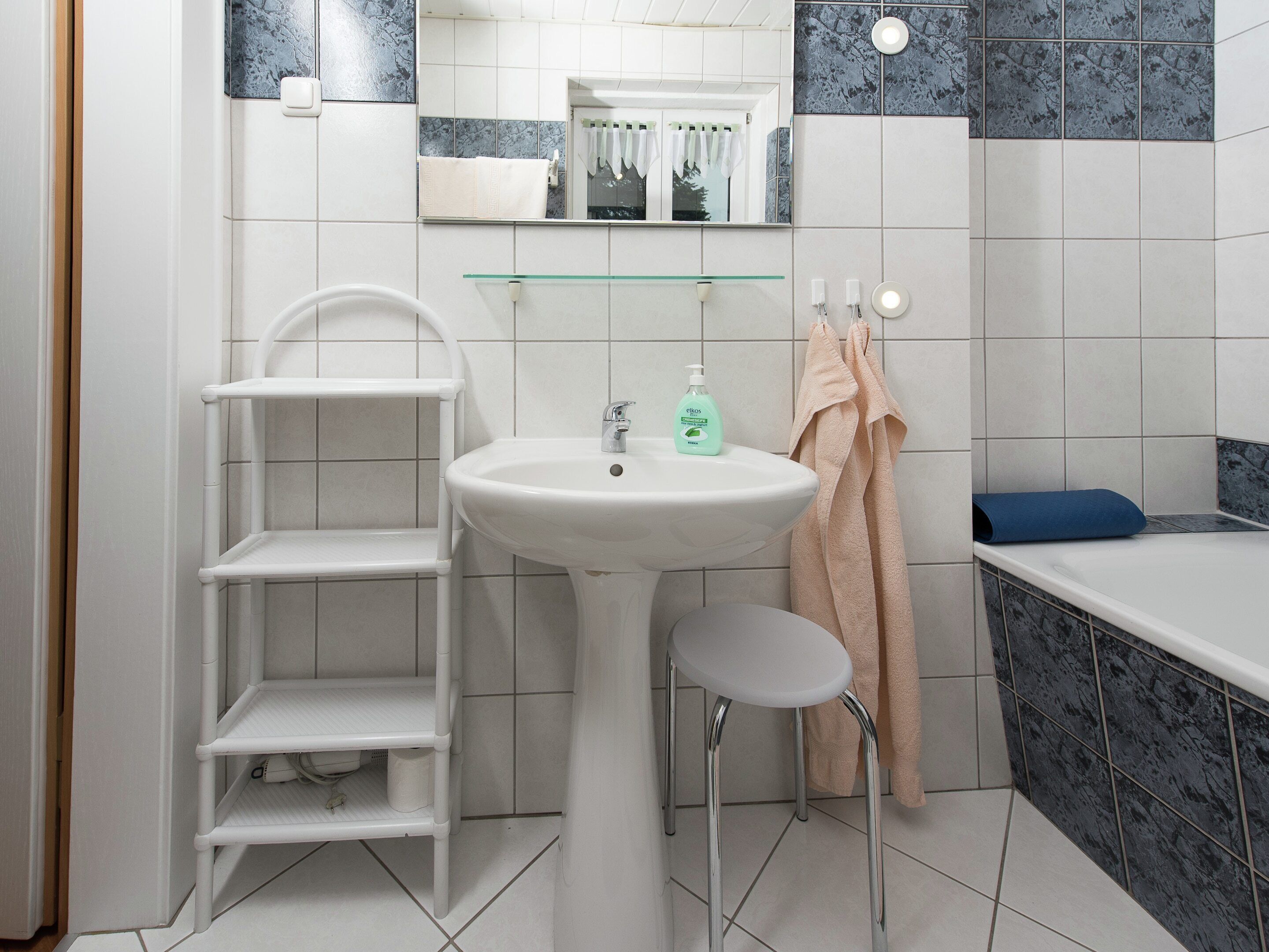 A bathroom with a white sink, a glass shelf, a white chair, a bathtub, and a mirror.