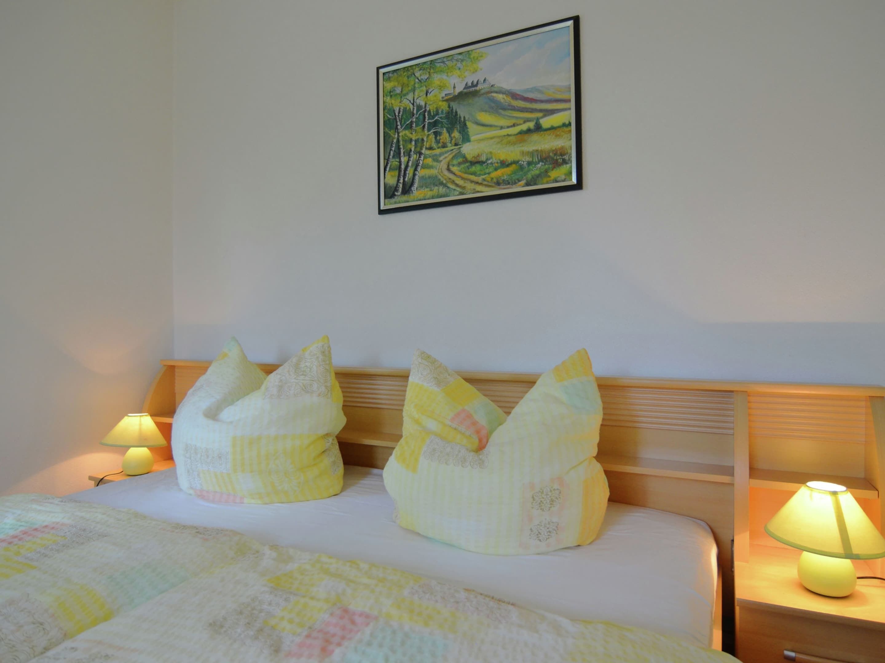 A neatly made bed with a quilt, two decorative pillows, a painting on the wall, and a bedside lamp.