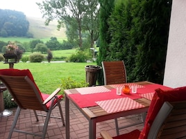 Un patio al aire libre con una mesa preparada para dos, sillas alrededor, vistas a un paisaje verde y una valla de madera.