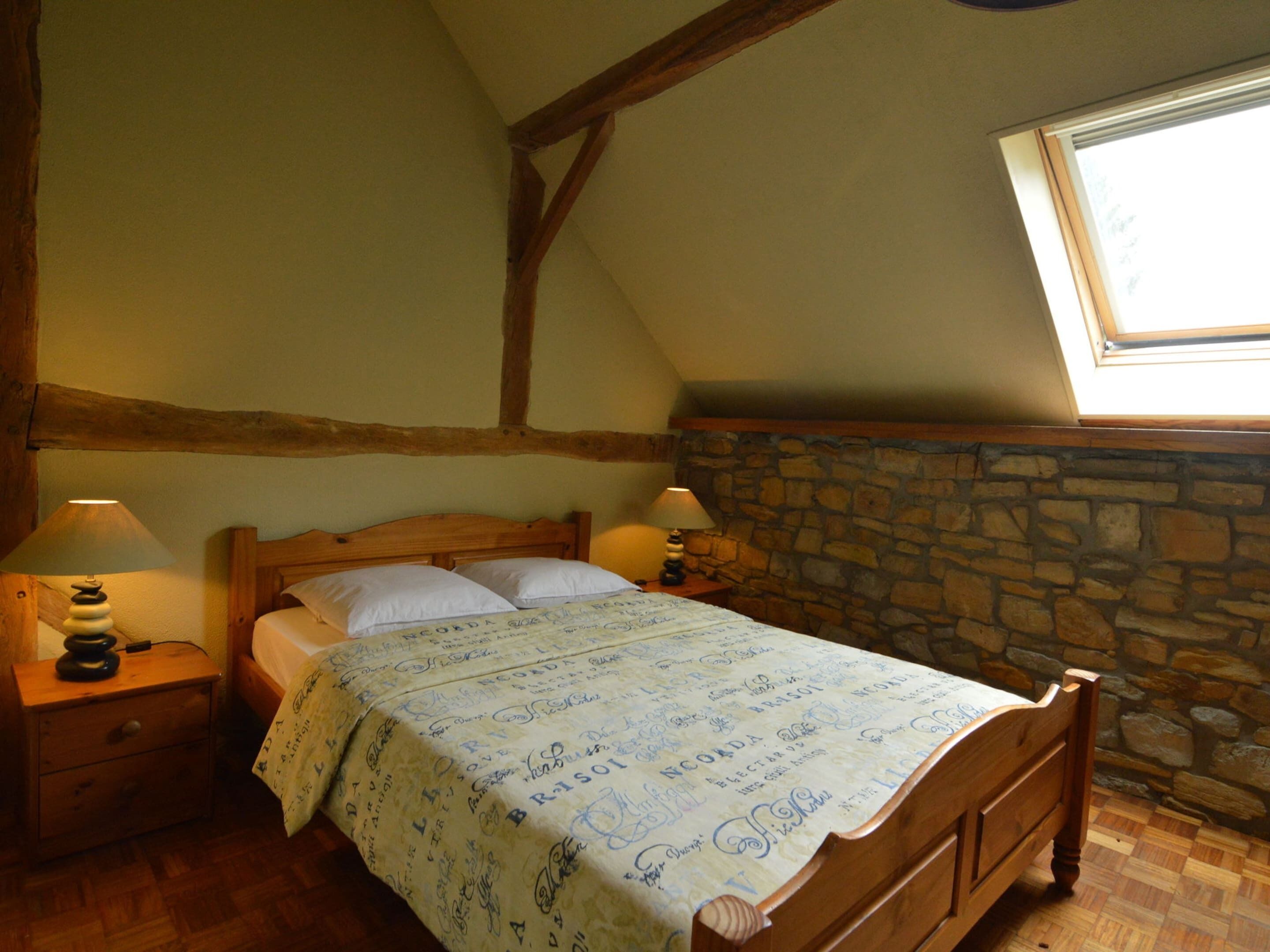A bedroom with a wooden bed, two bedside tables with lamps, a stone wall, and a skylight.