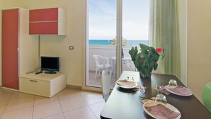 A room with a dining table set for two, a television on a cabinet, a sliding glass door leading to a balcony with a view of the sea, and a potted plant with a red flower. - Coastal Retreat in Riccione (Riccione)