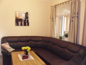 A living room with a dark brown sofa, a wooden coffee table, and a vase with yellow flowers. There is a painting on the wall and a window with curtains. - Bungalow Borgerende Nahe Strand & Reiten (Boergerende-Rethwisch)