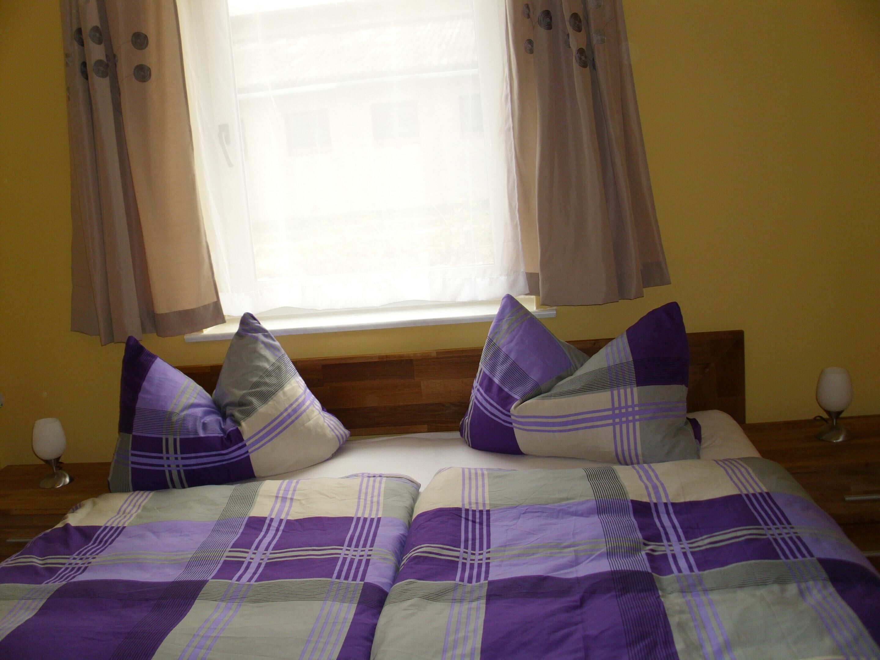 Ein Bett mit einer violett-weißen Karomattraze, zwei Kissen und einem Holz-Kopfteil.
