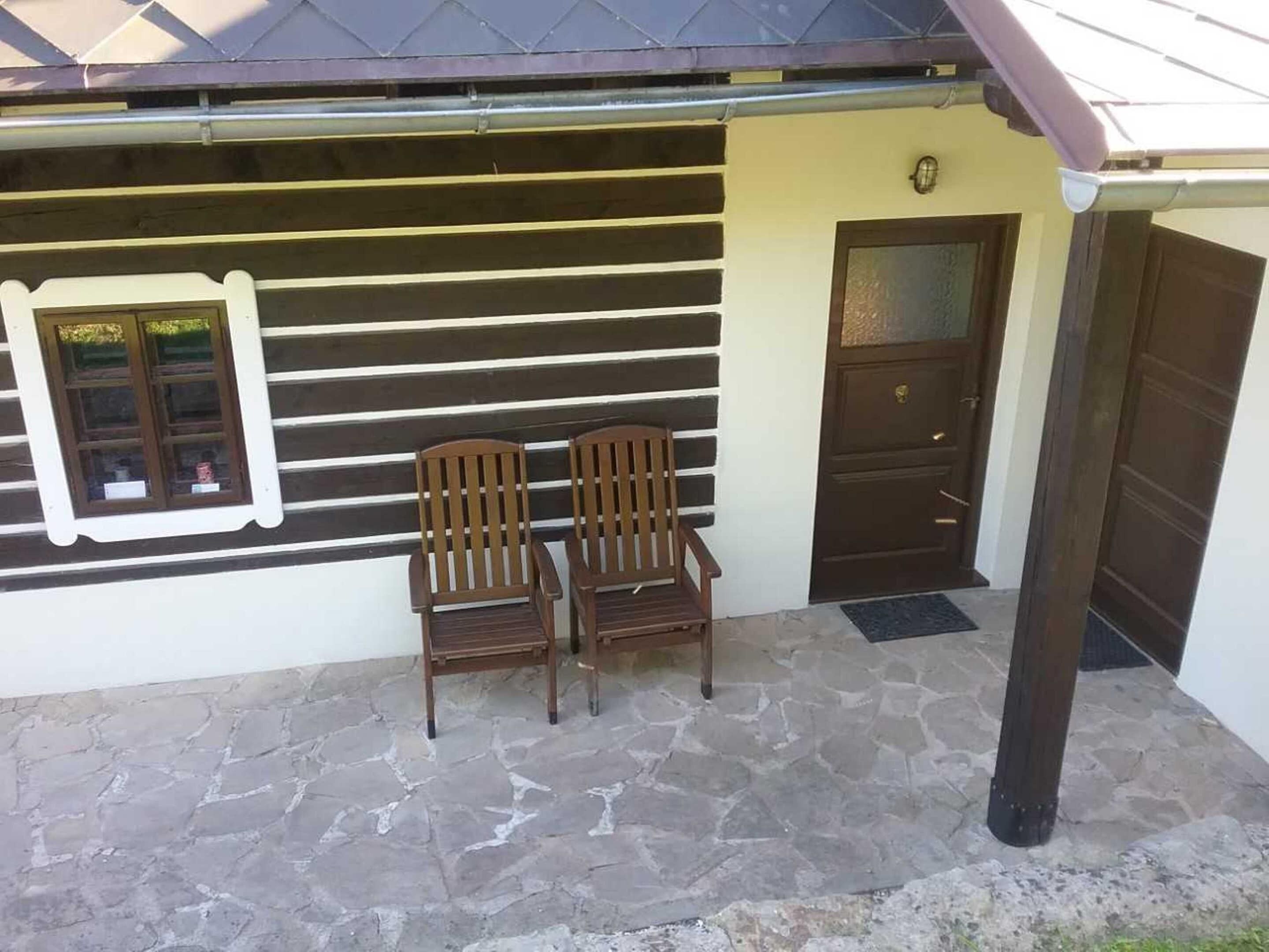 A building with a stone-paved entrance, two wooden chairs, a window with a view, and a door with a small sign above it.