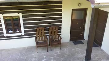 A building with a stone-paved entrance, two wooden chairs, a window with a view, and a door with a small sign above it.