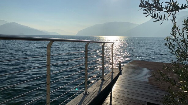 Une jetée en bois s’étendant sur des eaux calmes, agrémentée d’une rambarde en métal, d’où l’on peut admirer un paysage montagneux sous un ciel dégagé.