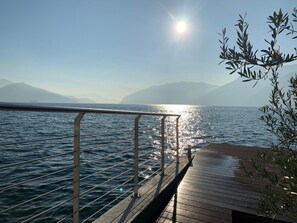 Une jetée en bois s’étendant sur des eaux calmes, agrémentée d’une rambarde en métal, d’où l’on peut admirer un paysage montagneux sous un ciel dégagé.