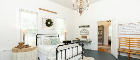 A bedroom with a bed, a chandelier, a desk, and a door leading to another room.