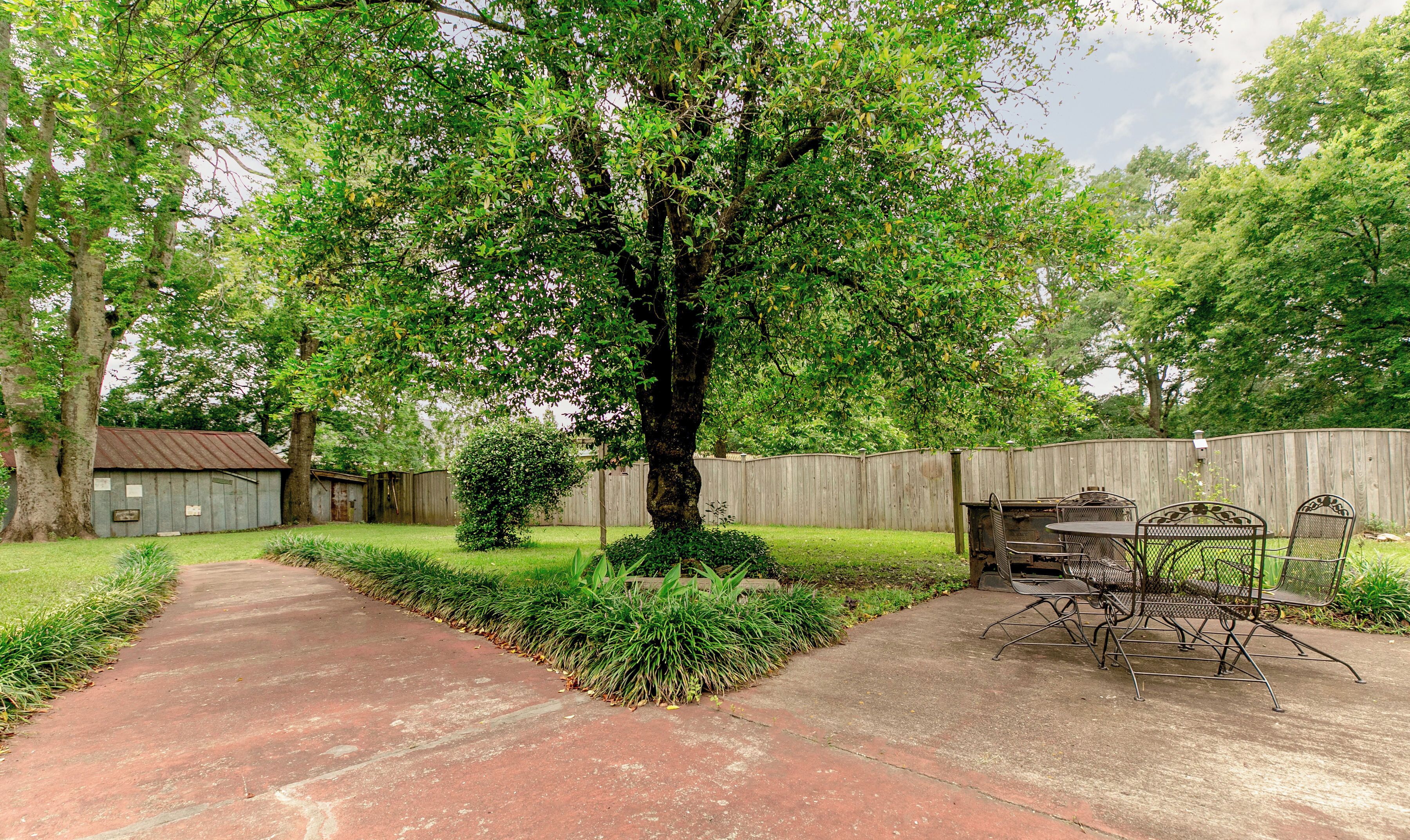 A backyard with a paved path, a metal table and chairs, a wooden shed, and a tall tree.