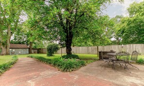 A backyard with a paved path, a metal table and chairs, a wooden shed, and a tall tree.