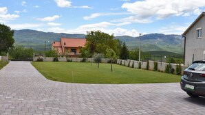 Une allée pavée mène à une maison avec un toit rouge, entourée d’une pelouse bien entretenue et avec une chaîne de montagnes en arrière-plan.