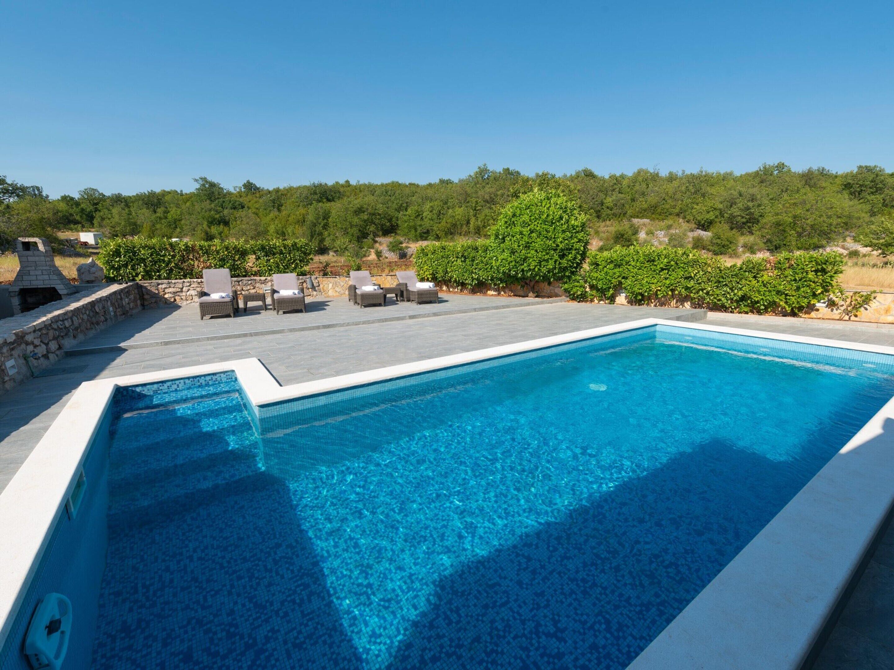 A rectangular swimming pool with clear blue water, surrounded by a stone deck and a stone wall. There are several lounge chairs and a barbecue grill visible in the background.