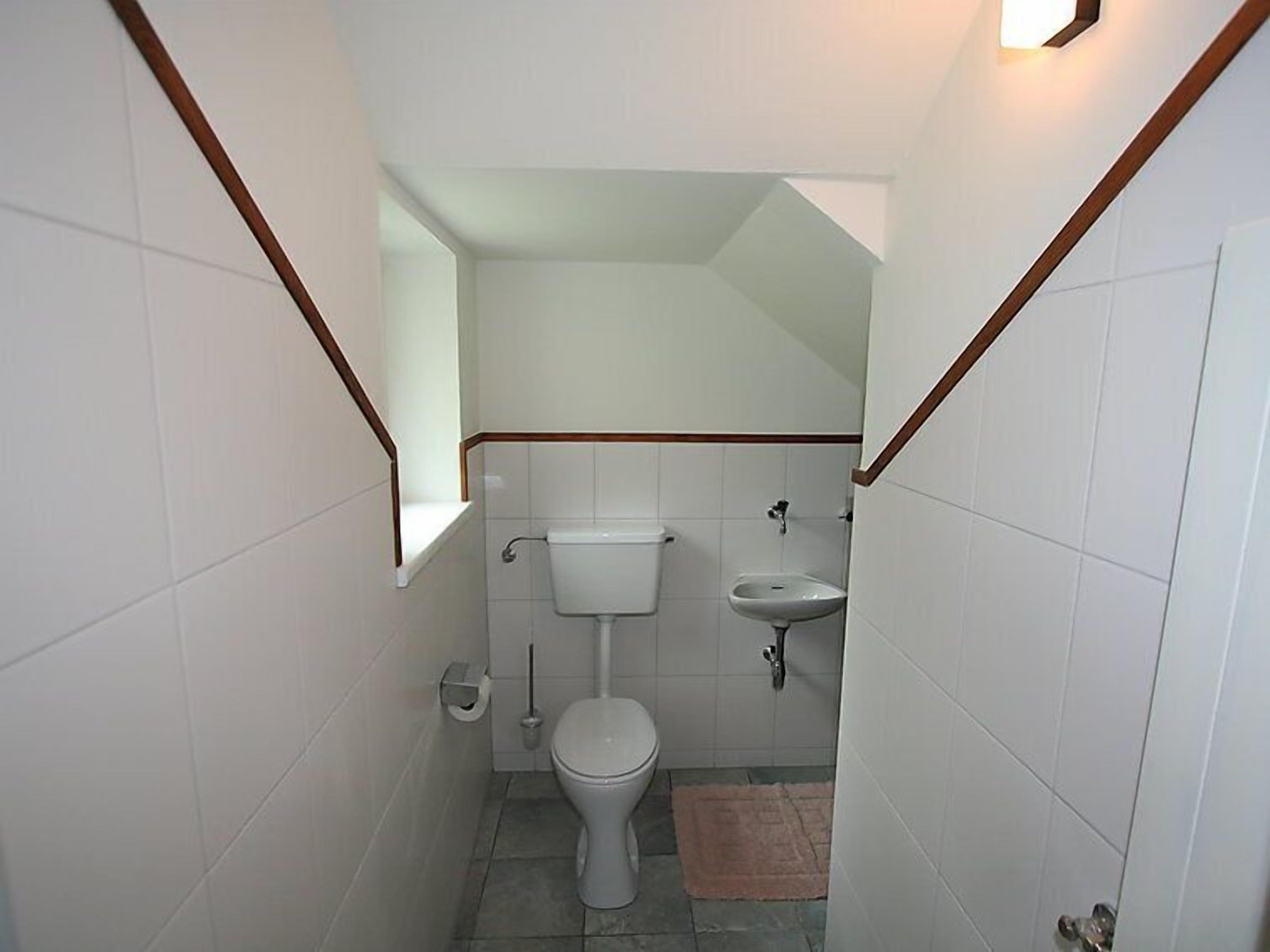 A small, narrow bathroom with white tiles, a toilet, a sink, and a window.