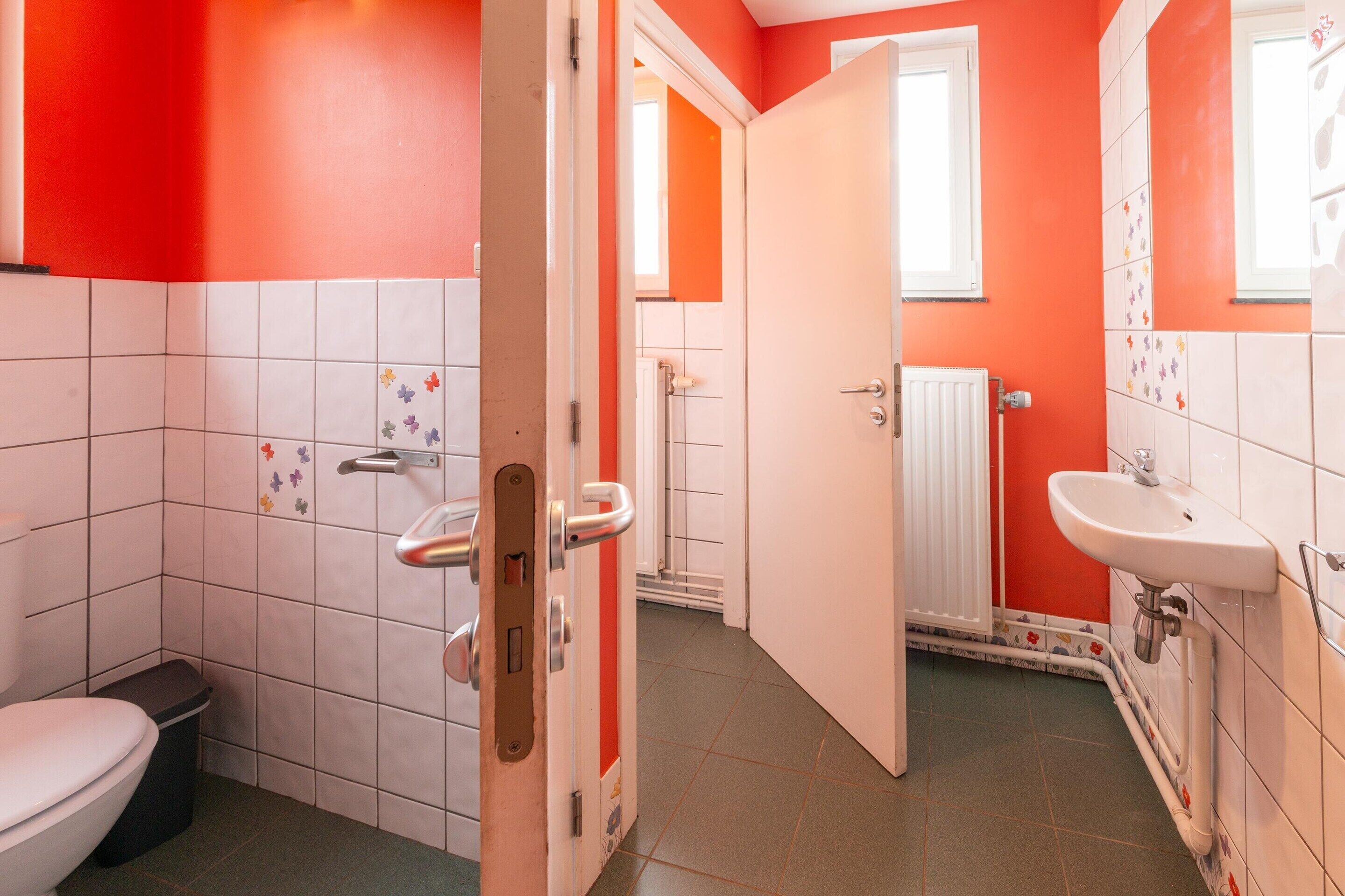 Ein Badezimmer mit roter Wand, weißen Fliesen, Toilette, Waschbecken und Duschbereich.