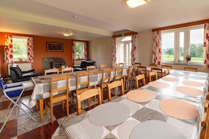 Een eetruimte met een lange tafel voor meerdere personen, omringd door stoelen, een open haard en uitzicht op de natuur door grote ramen.
