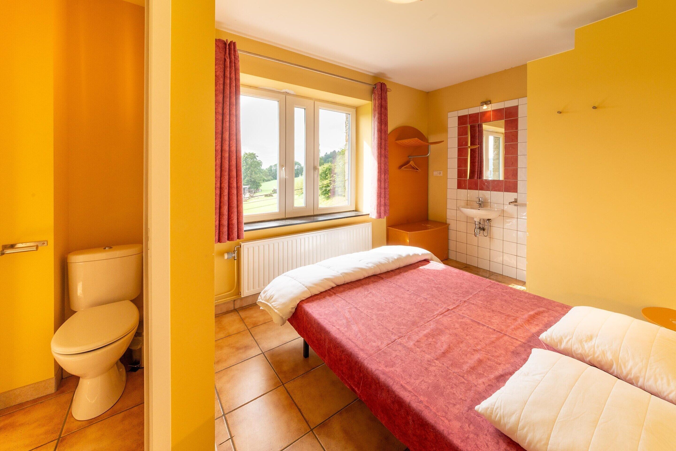A room with a bed, a window with curtains, a toilet, and a bathroom with a sink and mirror.