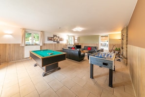 Een pooltafel, een tafelvoetbalspel en een bank in een goed verlichte kamer met houten wanden en een betegelde vloer.