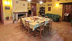 Un comedor con una mesa de madera dispuesta para cuatro personas, rodeada de sillas. Se ve una chimenea al fondo, y un sofá verde está colocado contra la pared.