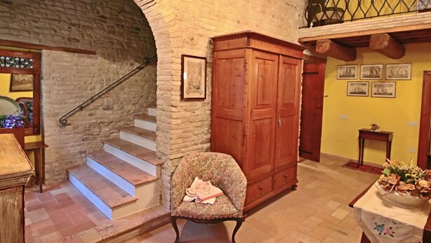 Una habitación con un arco de piedra, una escalera de madera, un armario de madera, un sillón con estampado floral y una mesa con un jarrón de flores.