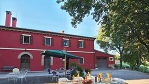 Un edificio rojo con una amplia mesa de patio dispuesta para comer, rodeada de sillas blancas y una sombrilla que proporciona sombra.
