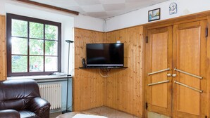 Een woonkamer met een leren bank, een houten tafel met een witte tafelkleed, een aan een houten wand bevestigde televisie en een grote houten garderobe met twee deuren.