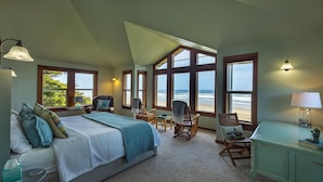 Un dormitorio con una cama grande, dos mecedoras, un escritorio y varias ventanas con vistas al océano.