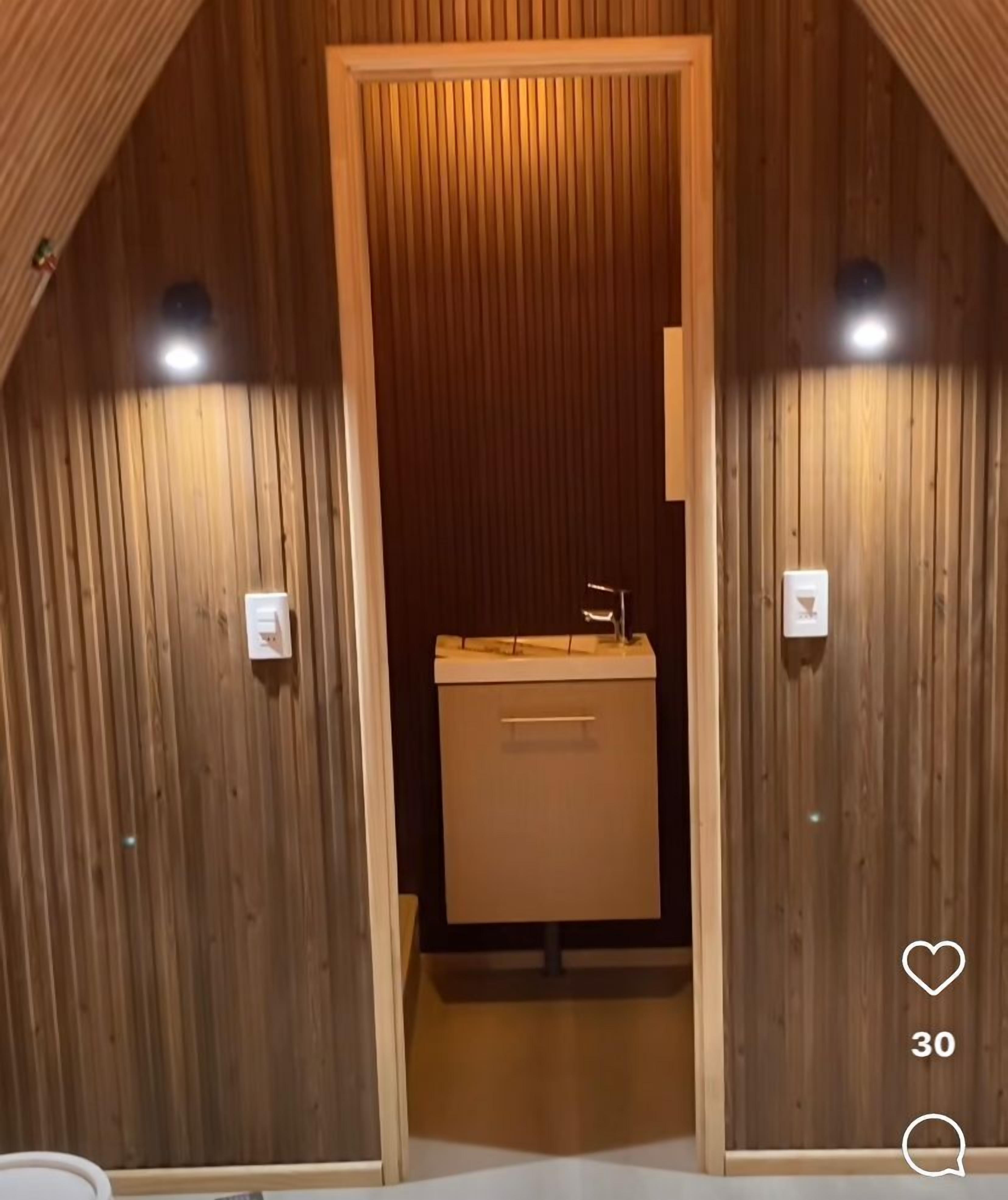 A wooden-paneled room with a central door leading to a bathroom. There are two wall-mounted light fixtures on either side of the door.