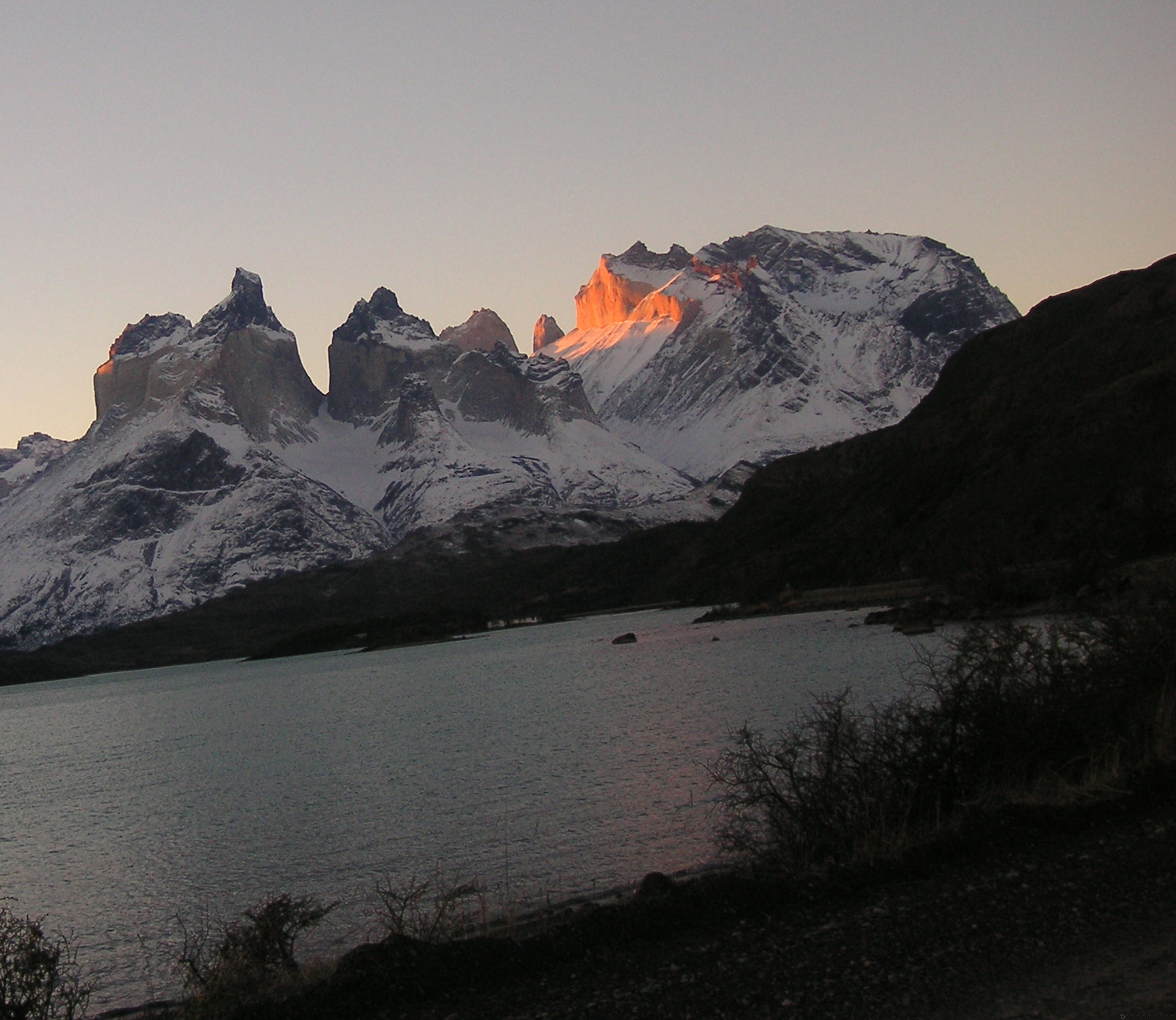 A mountain range with snow-covered peaks and a calm lake in the foreground.
