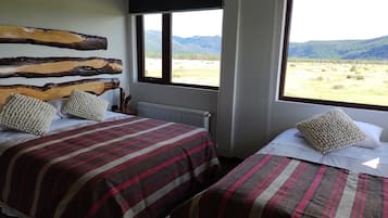 A room with two beds, each with striped bedding and decorative pillows, a wooden headboard, and a view of a mountainous landscape through large windows.