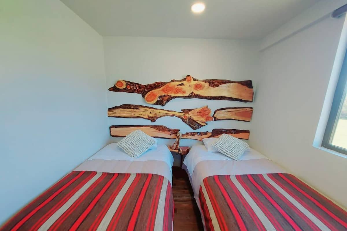 Ein Zimmer mit zwei Betten, jeweils mit gestreifter Bettwäsche, einem Holzkopfende mit aufgemaltem Dekor und einem Fenster auf der rechten Seite.