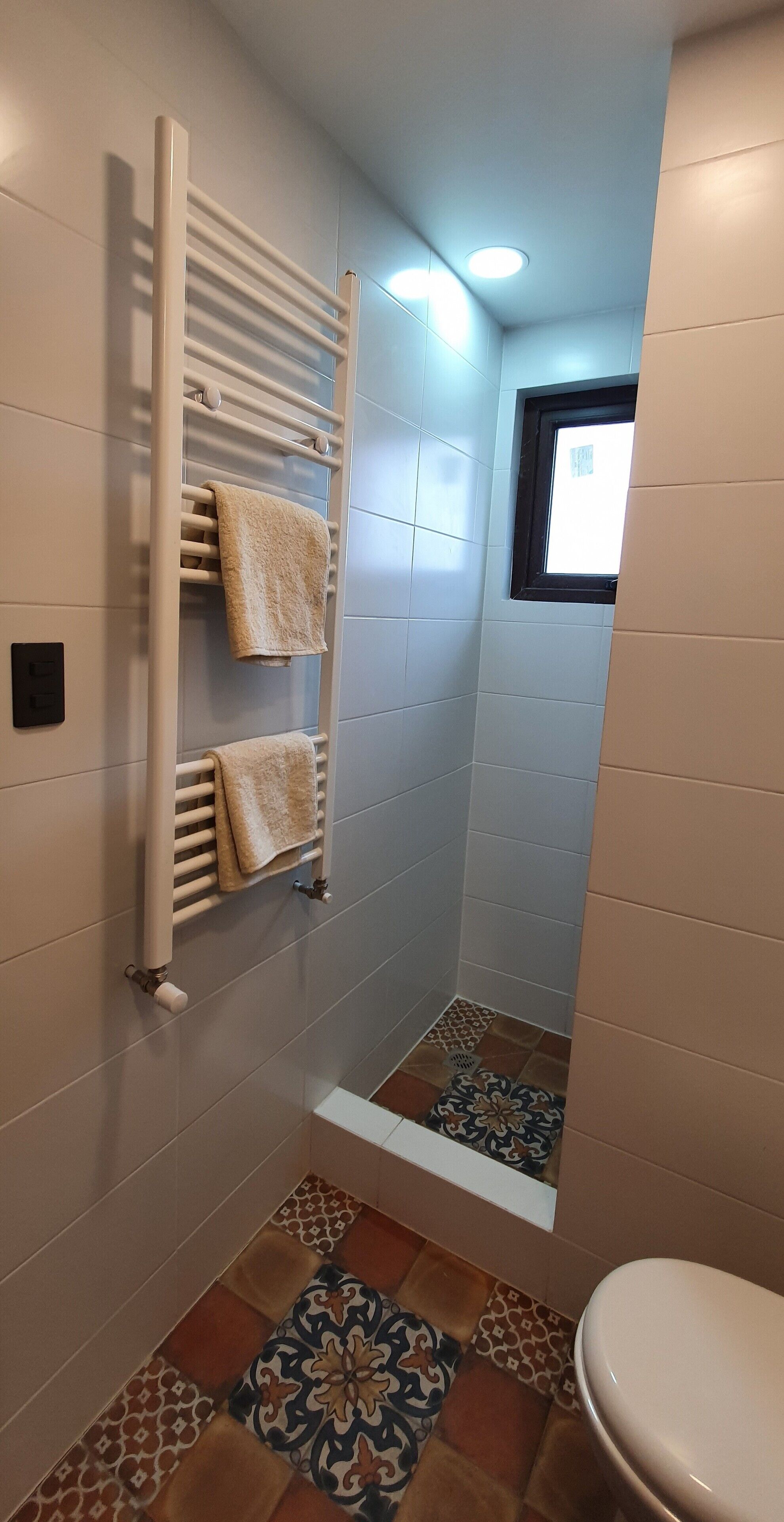 Un baño pequeño con ducha, un tendedero para toallas con dos toallas, una ventana y baldosas estampadas en el suelo.