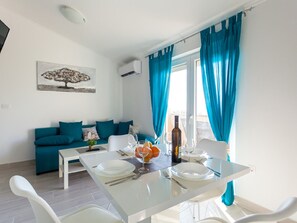 Un salon moderne avec un coin repas. On y trouve un canapé avec des coussins turquoise, une petite table avec un plateau en verre et une table à manger dressée avec des assiettes, des verres et une bouteille. Un tableau représentant un arbre est accroché au mur, et il y a une fenêtre avec des rideaux bleus.