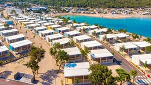 En rad moderna, vita husvagnar med stora fönster, placerade längs en väg som går intill stranden, med utsikt över havet och en skogbeklädd kulle i bakgrunden.