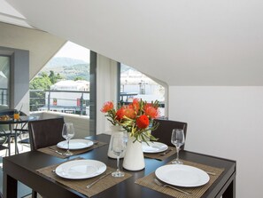 A dining table set with plates, cutlery, and a vase of flowers, with a view of buildings and hills through large windows. - Topfloor Comfortable Luxury Apartment With Private Balcony,free Garage and Wi-fi (Dubrovnik)
