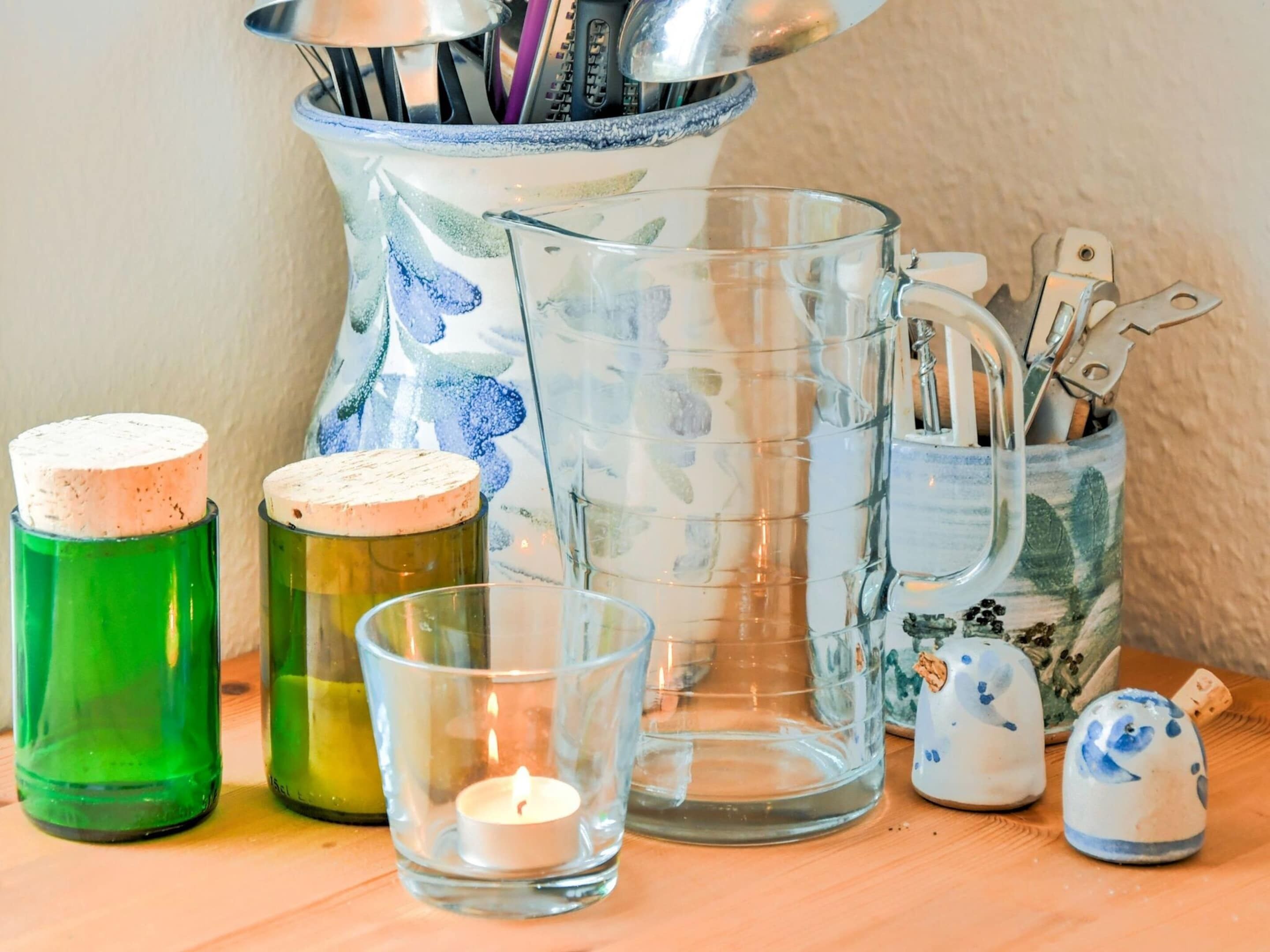 Verschiedene Glasbehälter, darunter eine grüne Flasche, ein transparenter Kerzenhalter und ein dekorativer Krug mit Besteck.