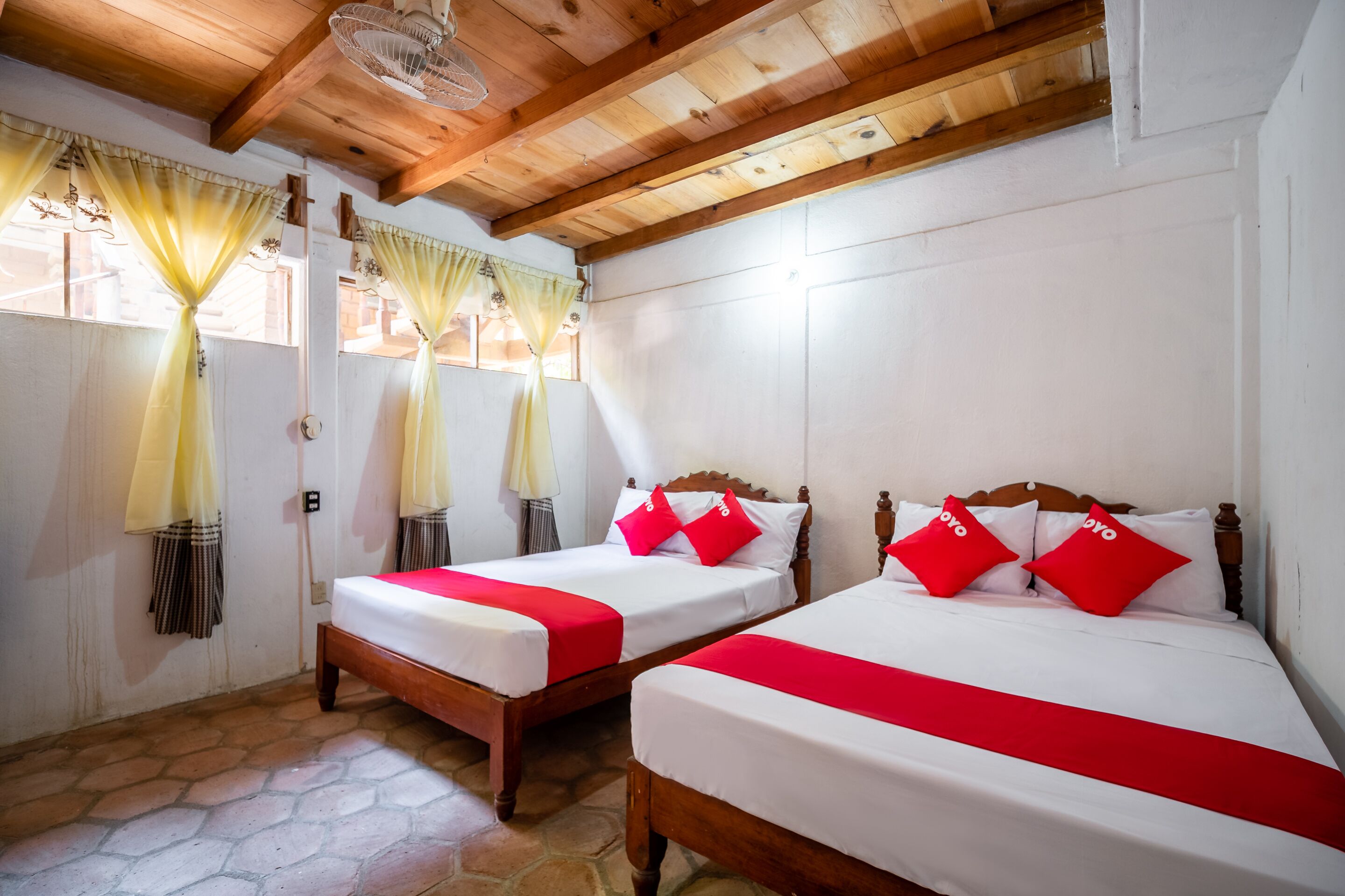 Una habitación con dos camas de madera, cada una con ropa blanca y detalles rojos, bajo un techo de madera con vigas a la vista.
