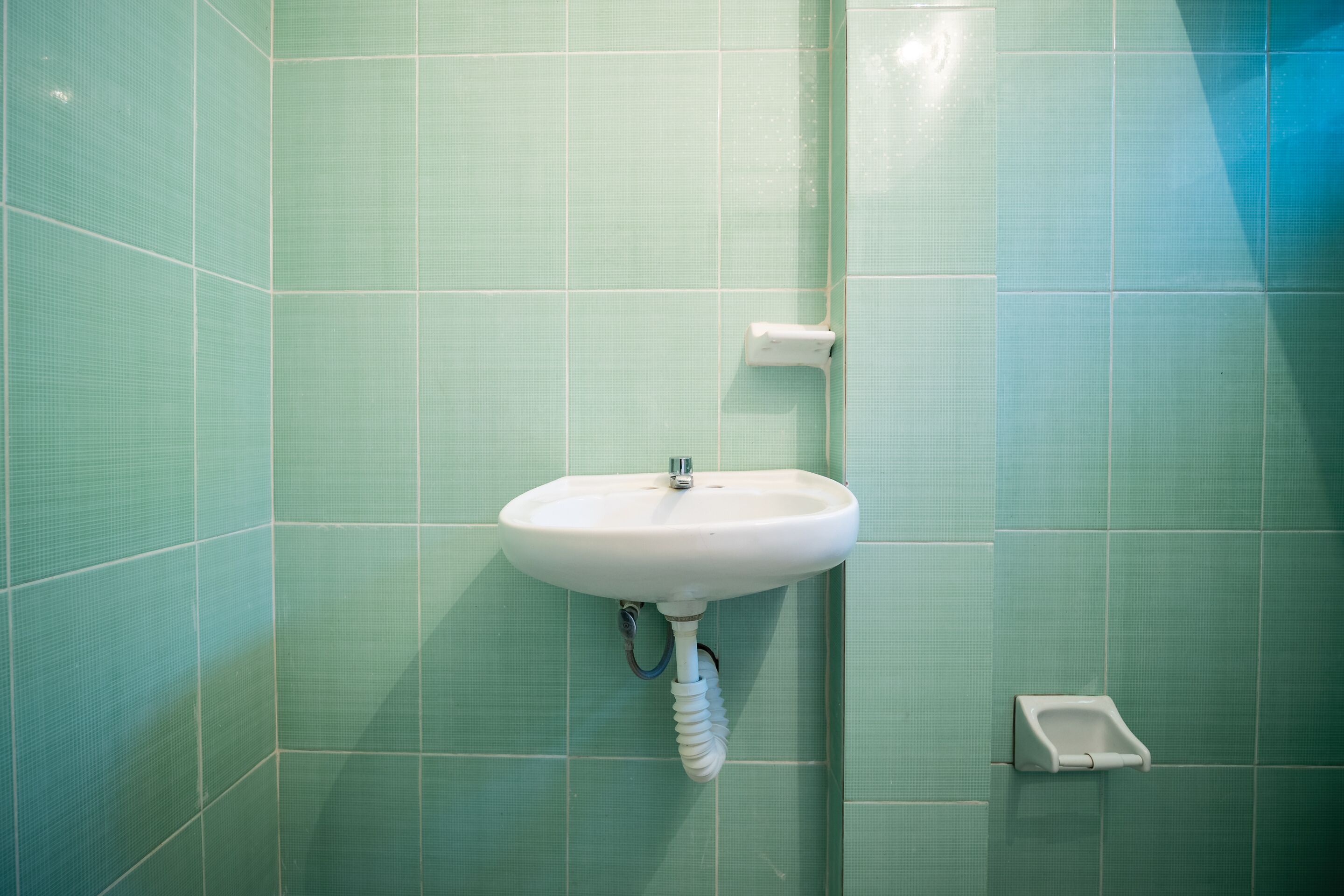 Une salle de bain avec un lavabo blanc fixé sur un mur recouvert de carreaux verts, un sèche-mains et une petite poubelle.