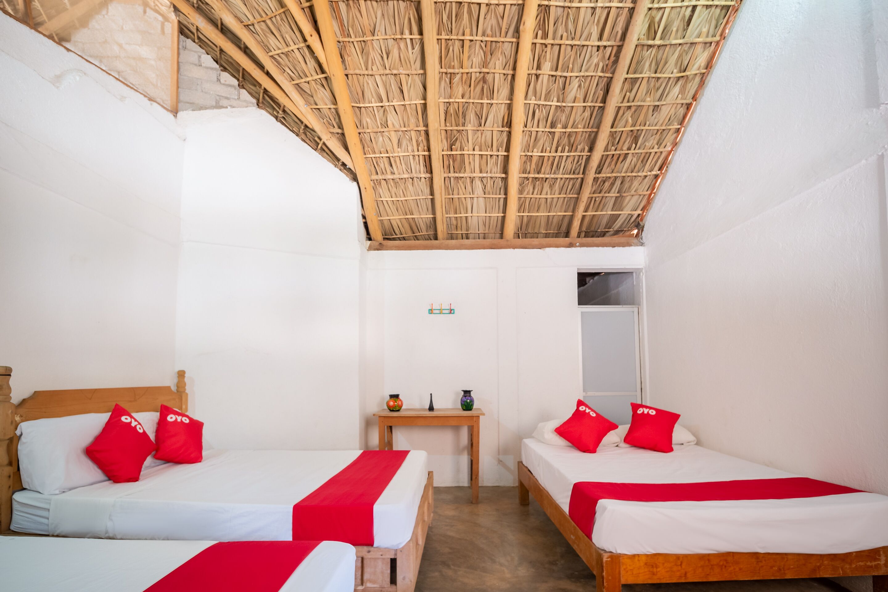 A room with two beds, each with red pillows and a white bedspread. There is a small wooden table between the beds with a vase and a candle. The room has a thatched roof ceiling and white walls.